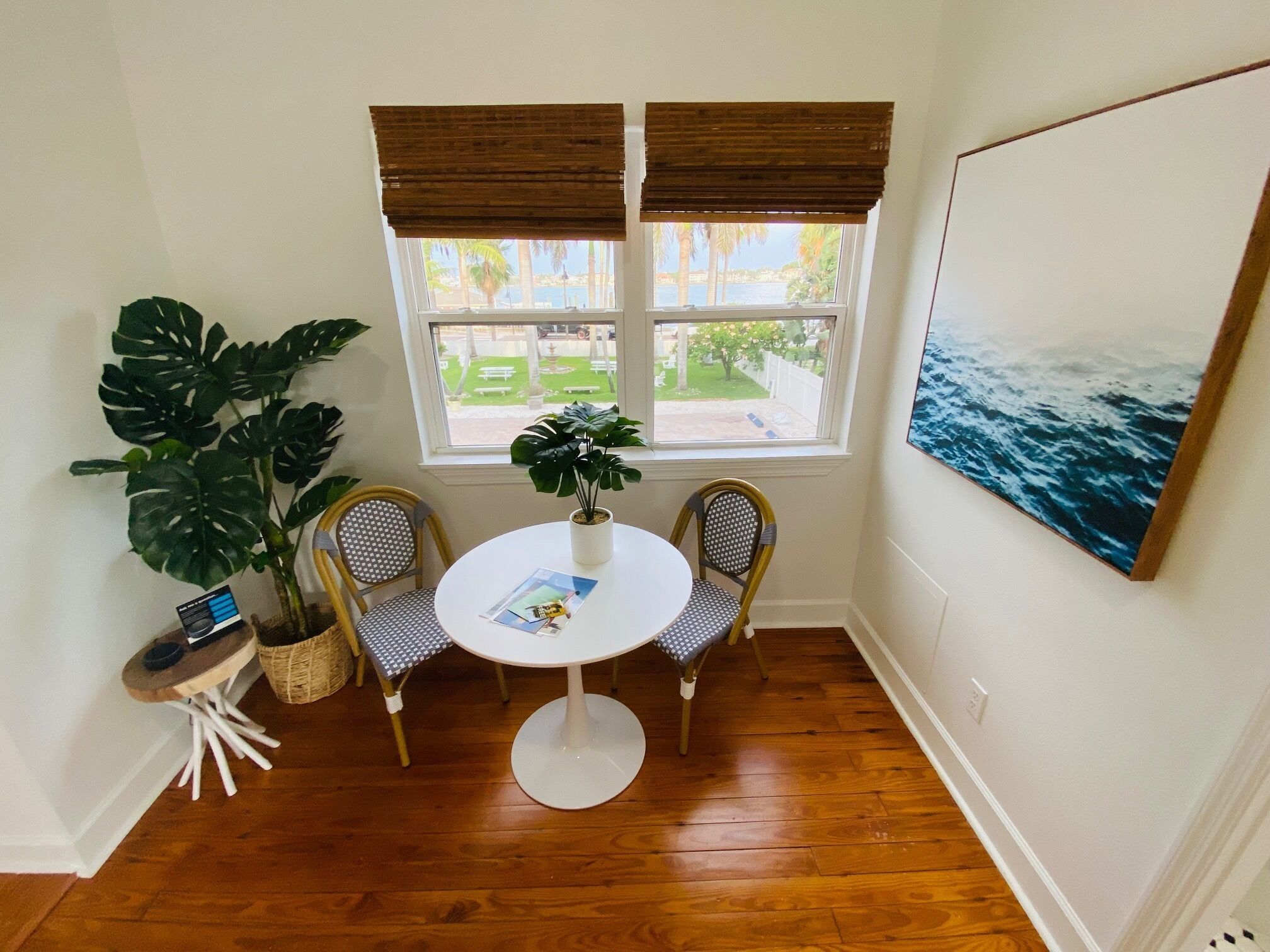 Una habitación con piso de madera, una mesa redonda con dos sillas, una planta en maceta, una mesita auxiliar y un cuadro en la pared.