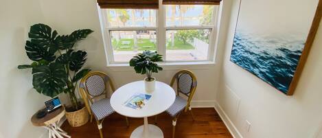 Una habitación con piso de madera, una mesa redonda con dos sillas, una planta en maceta, una mesita auxiliar y un cuadro en la pared.