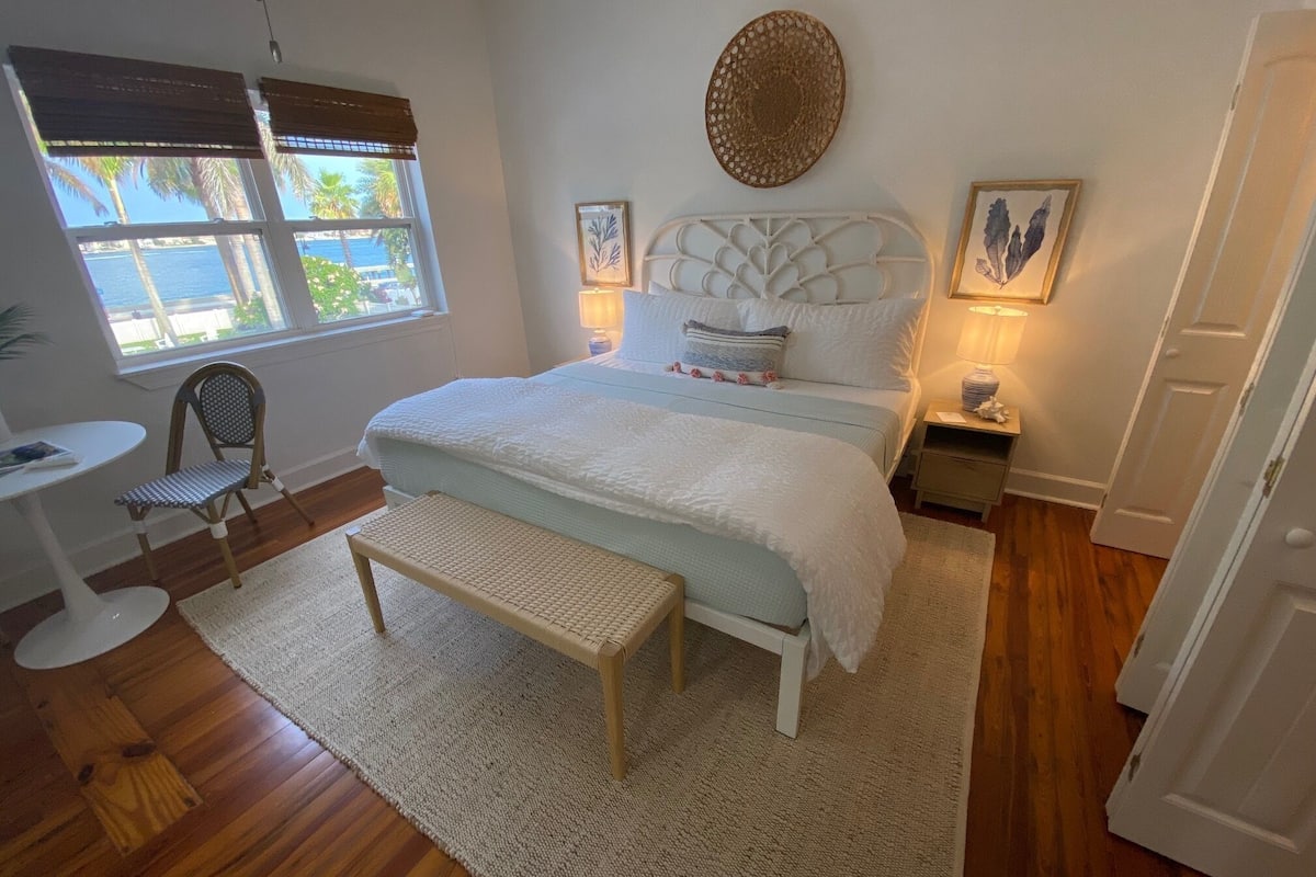 Un dormitorio con una cama grande, una mesita de noche, una silla, una ventana con persianas y vistas a palmeras.