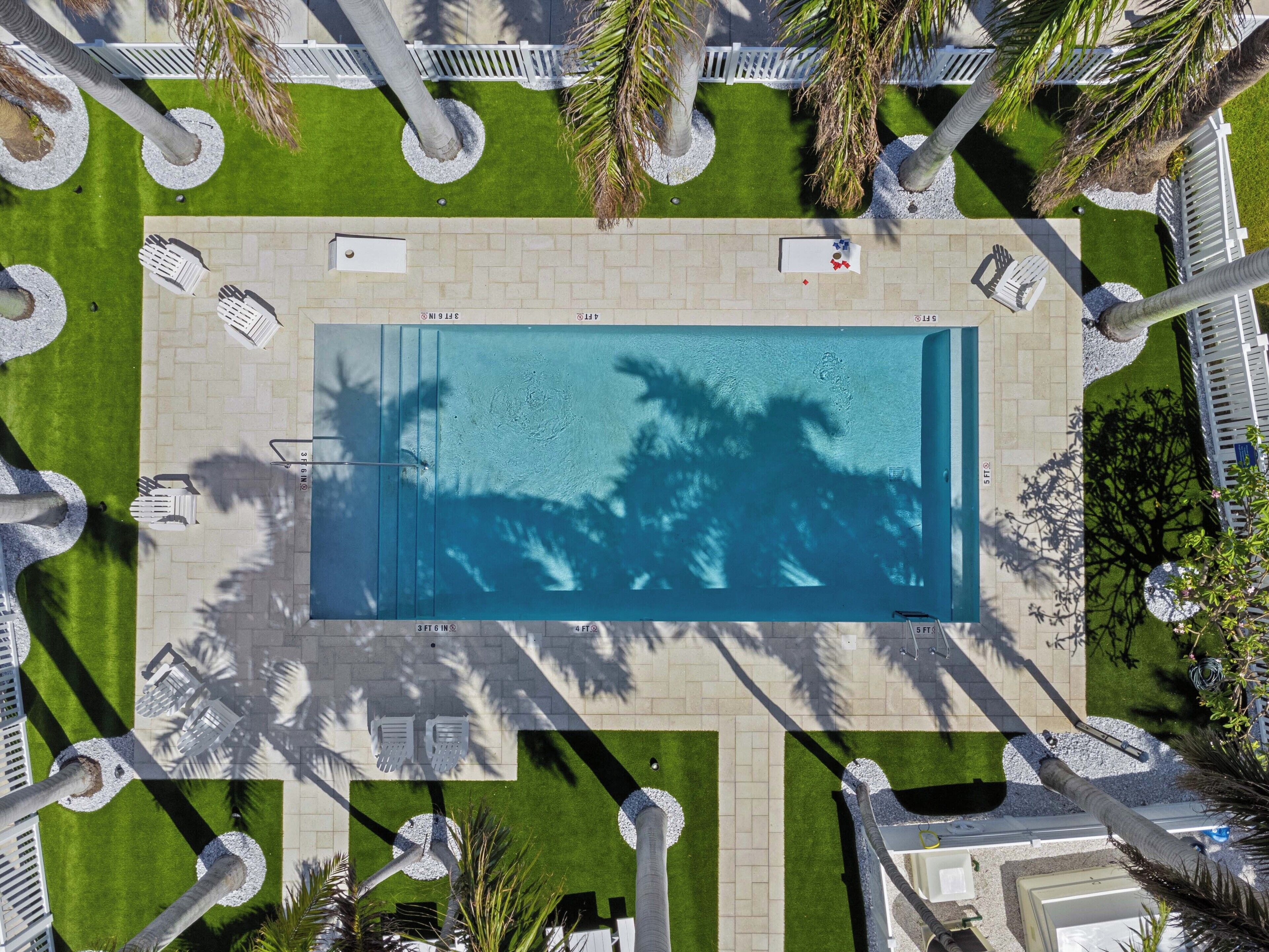 Vista aérea de una piscina rectangular rodeada de vallas blancas, palmeras y un camino pavimentado.