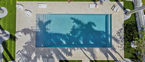 Vista aérea de una piscina rectangular rodeada de vallas blancas, palmeras y un camino pavimentado.