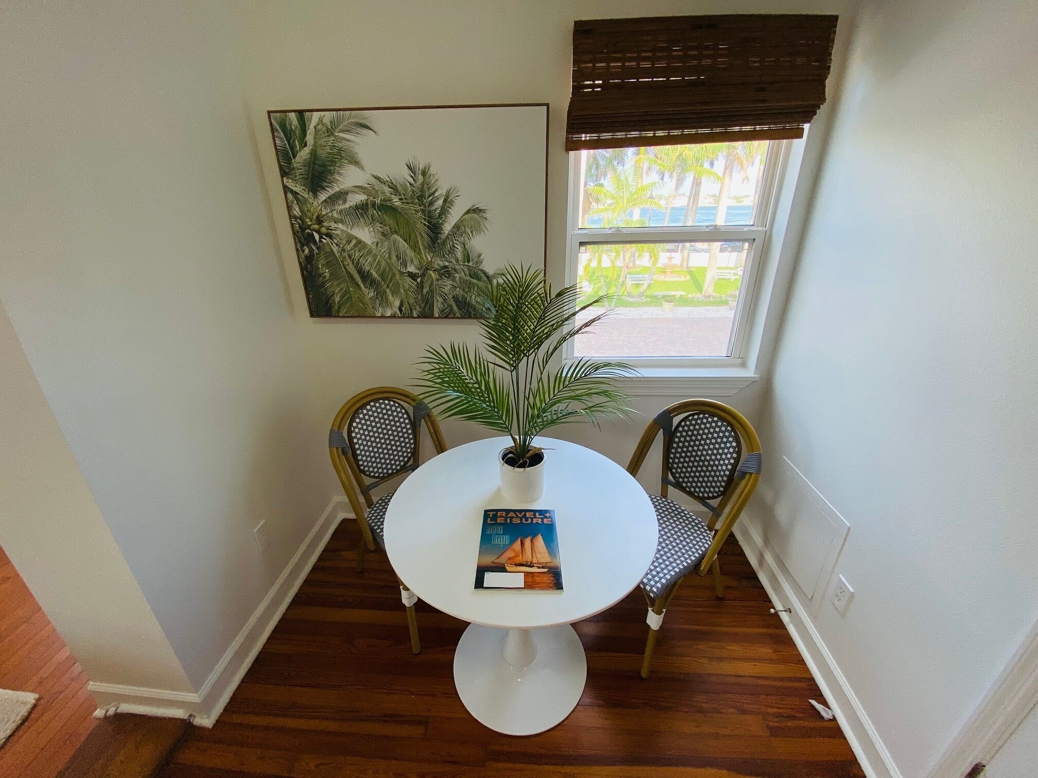 A room with a round table, two chairs, a potted plant, a framed picture, and a window with a view of palm trees and a building.