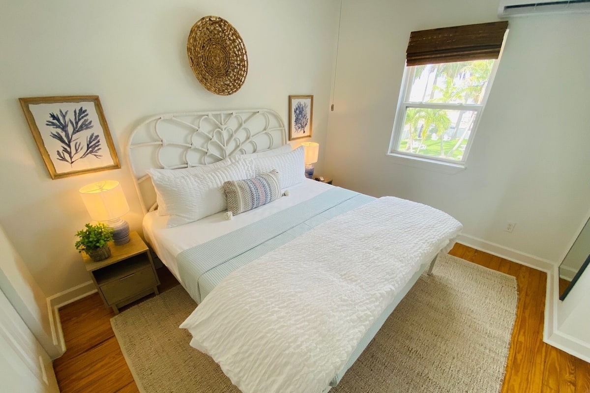 Un dormitorio con cama, mesitas de noche, lámpara, planta y ventana con vista a follaje.