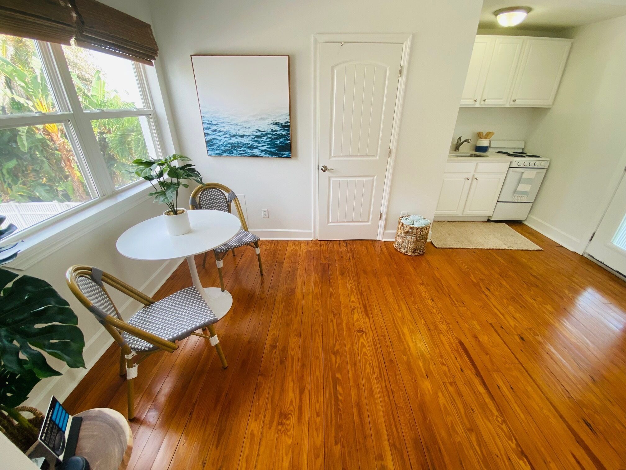 Una habitación con piso de madera, una mesa de comedor con dos sillas, una planta en maceta, una puerta blanca y una cocina con gabinetes y electrodomésticos.