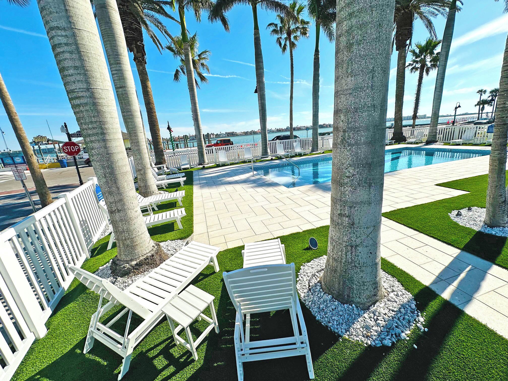 Zona junto a la piscina con sillas reclinables blancas, palmeras y vista al agua.