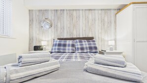 A bedroom with a wooden wall panel, a round mirror, a bed with plaid bedding, two bedside tables with lamps, and a door. - Castle Cove - 1 Bedroom Apartment - Tenby (Tenby)