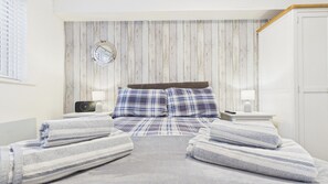 A bedroom with a wooden wall panel, a round mirror, a bed with plaid bedding, two bedside tables with lamps, and a door.