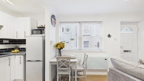 Eine kleine Küche mit weißen Schränken, einer Mikrowelle und einem Kühlschrank. Ein Esstisch mit Stühlen und eine Vase mit Sonnenblumen sind vorhanden. Ein Fenster mit Jalousien ist sichtbar, und eine weiße Tür befindet sich rechts.