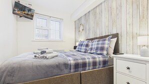 Un dormitorio con cama, mesita de noche, lámpara, televisor montado en la pared y ventana con persianas.