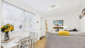 A modern living room with a grey sofa, a dining table with chairs, a vase of sunflowers, and a painting of a beach scene. - Castle Cove - 1 Bedroom Apartment - Tenby (Tenby)