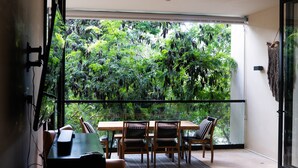Un balcon avec une table et des chaises en bois, une vue sur une végétation luxuriante à travers une porte vitrée et une décoration murale en macramé.