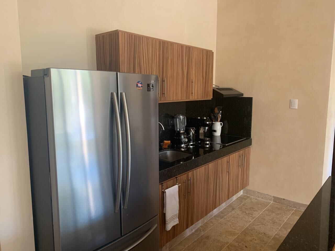 A kitchen with a stainless steel refrigerator, wooden cabinets, a black countertop, and a tiled floor.