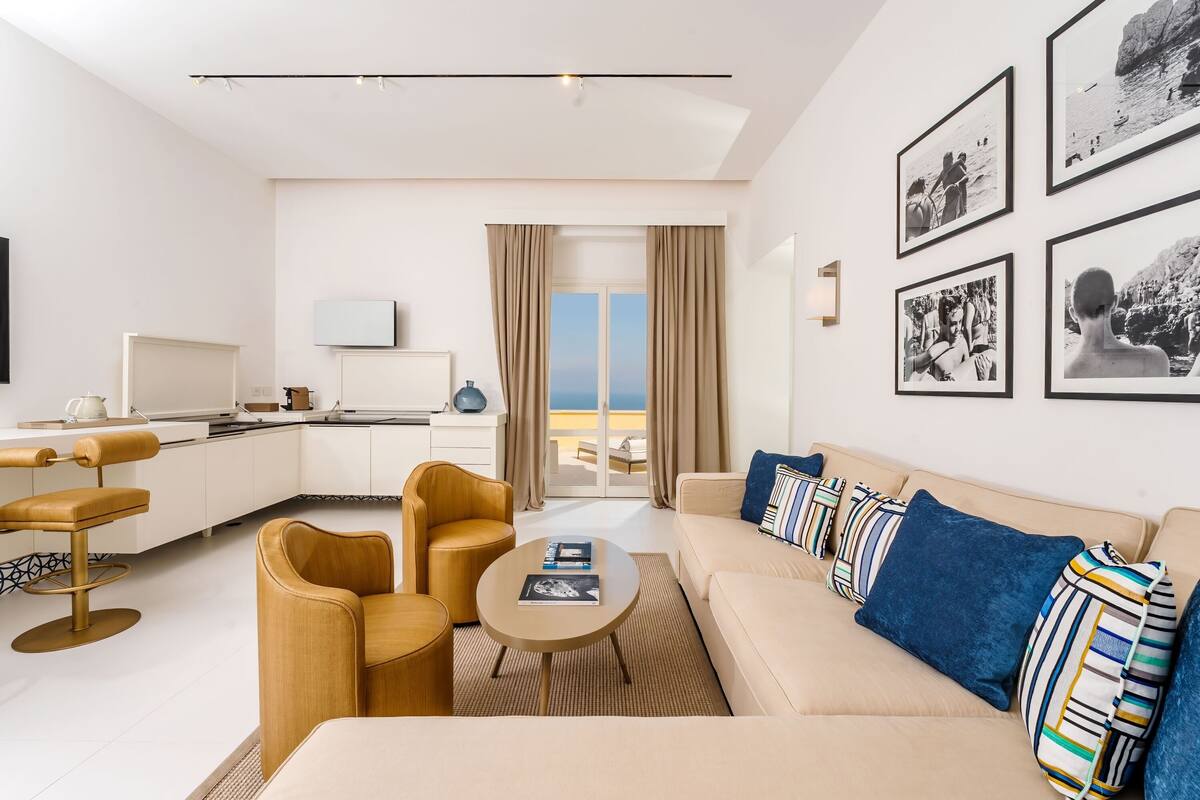 A modern hotel room with a sofa, coffee table, and a dining area with chairs and a table.