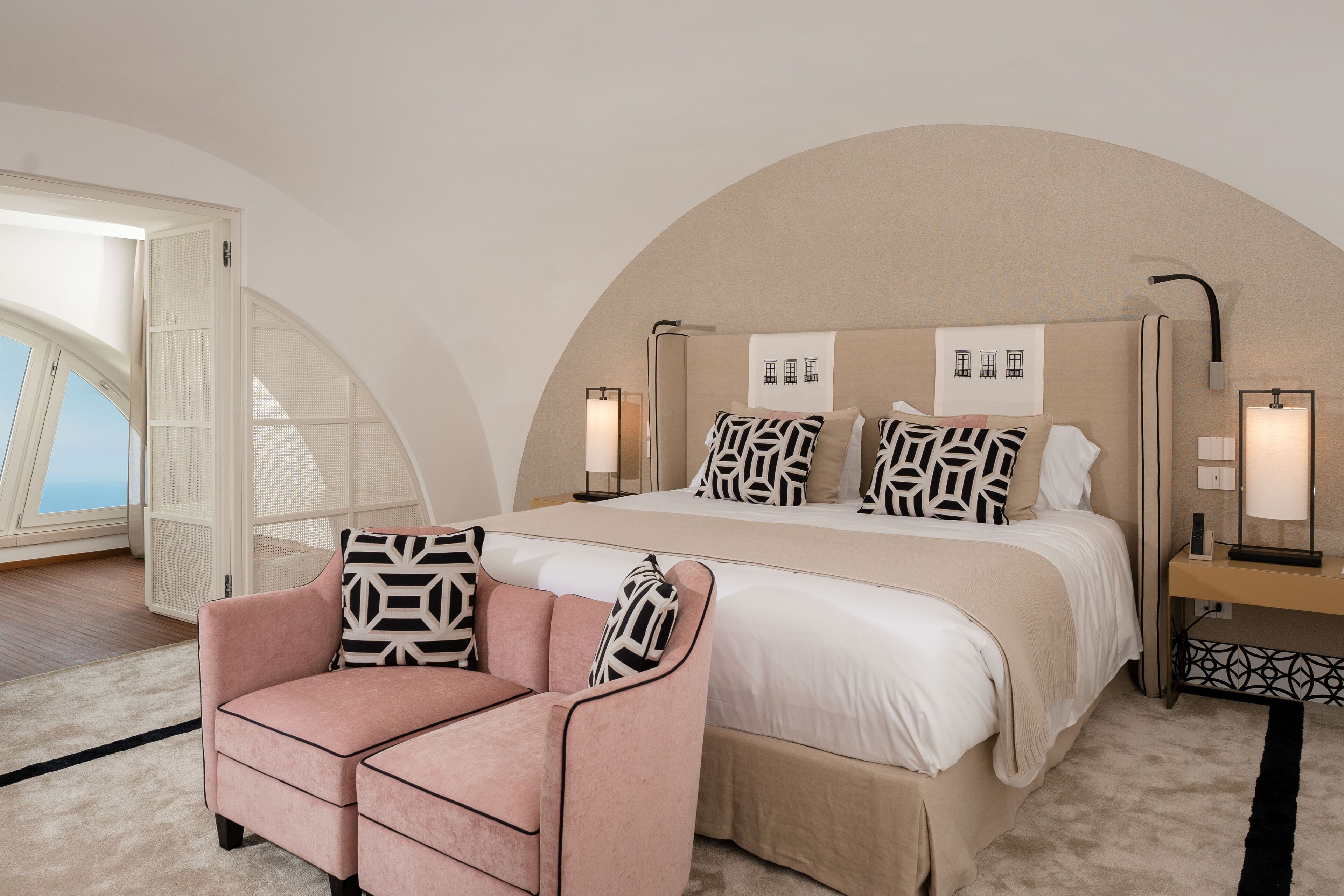 A bedroom with a large bed, a pink armchair, and a view of the sea through a door.