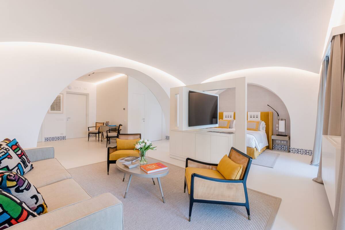 A modern hotel room with a living area featuring a sofa, armchair, and a small coffee table. There is a flat-screen TV mounted on the wall, a bed with a nightstand, and a dining area with a table and chairs.