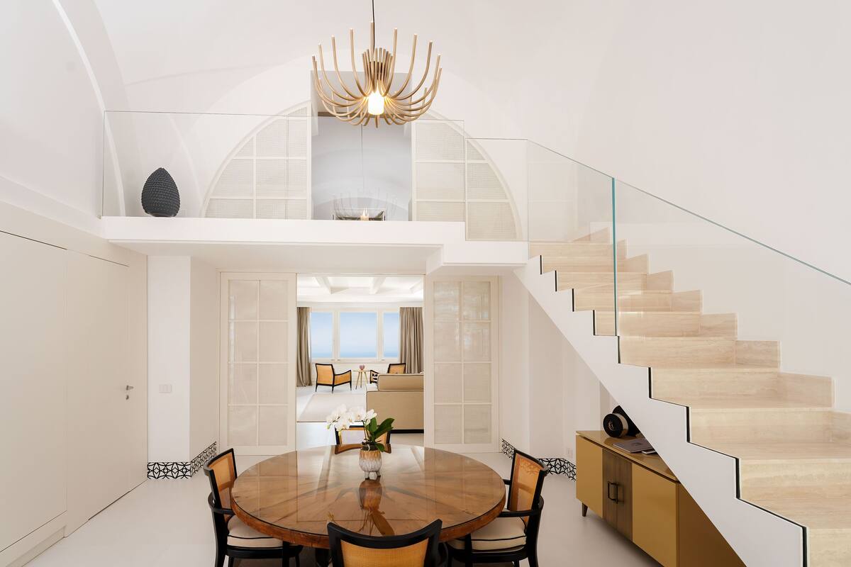 A modern dining area with a round table, chairs, and a chandelier. A staircase with glass railings leads to an upper level.