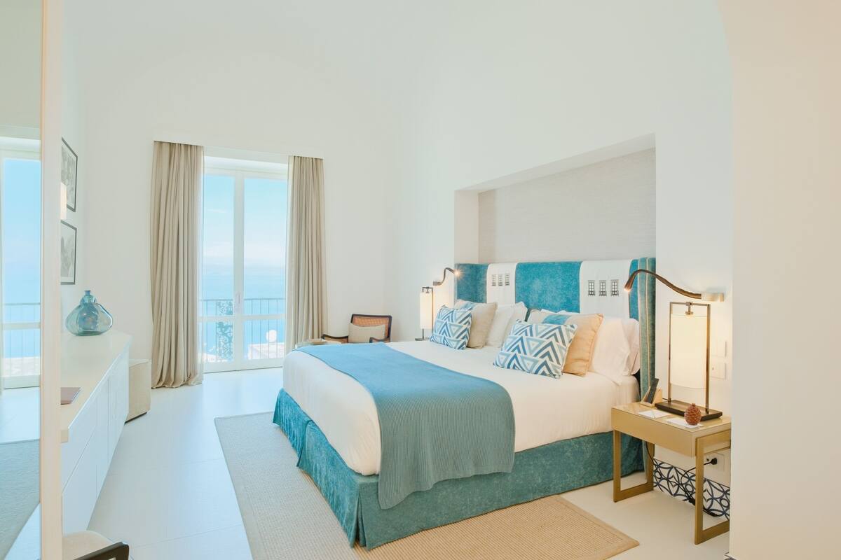 A modern hotel room with a large bed, a bedside table with a lamp, a view of the outdoors, and a window with curtains.