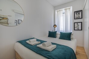 A neatly made bed with teal bedding and white towels, a round mirror on the wall, and two framed pictures on the wall. - Studio with a patio (Lisboa)