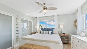 Une chambre à coucher comprenant un lit, un ventilateur de plafond, une table de chevet, une commode, une fenêtre avec vue et un banc en osier.