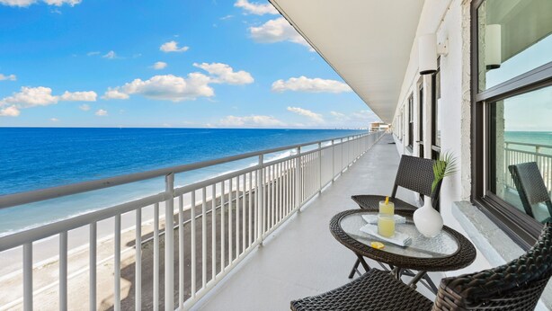 Un balcon avec vue sur l’océan, aménagé avec des fauteuils et une table en osier, et un parasol blanc pour offrir de l’ombre.