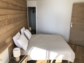 A bedroom with a wooden headboard, a bed with white bedding, a bedside table with a lamp, and a door leading to another room.
