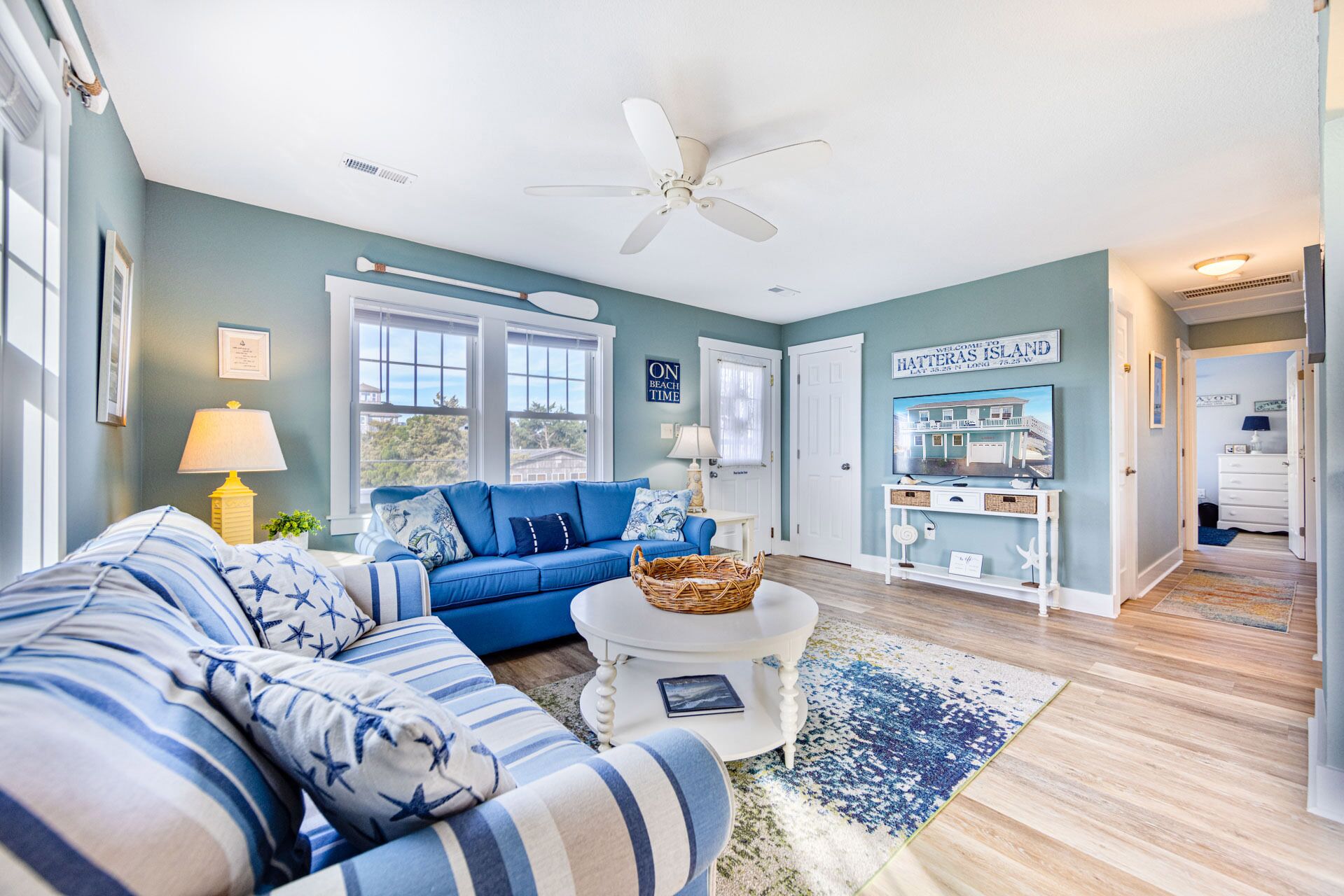 Un salon avec un canapé bleu, une table basse blanche et un ventilateur de plafond. Une grande fenêtre offre une vue sur l’extérieur.