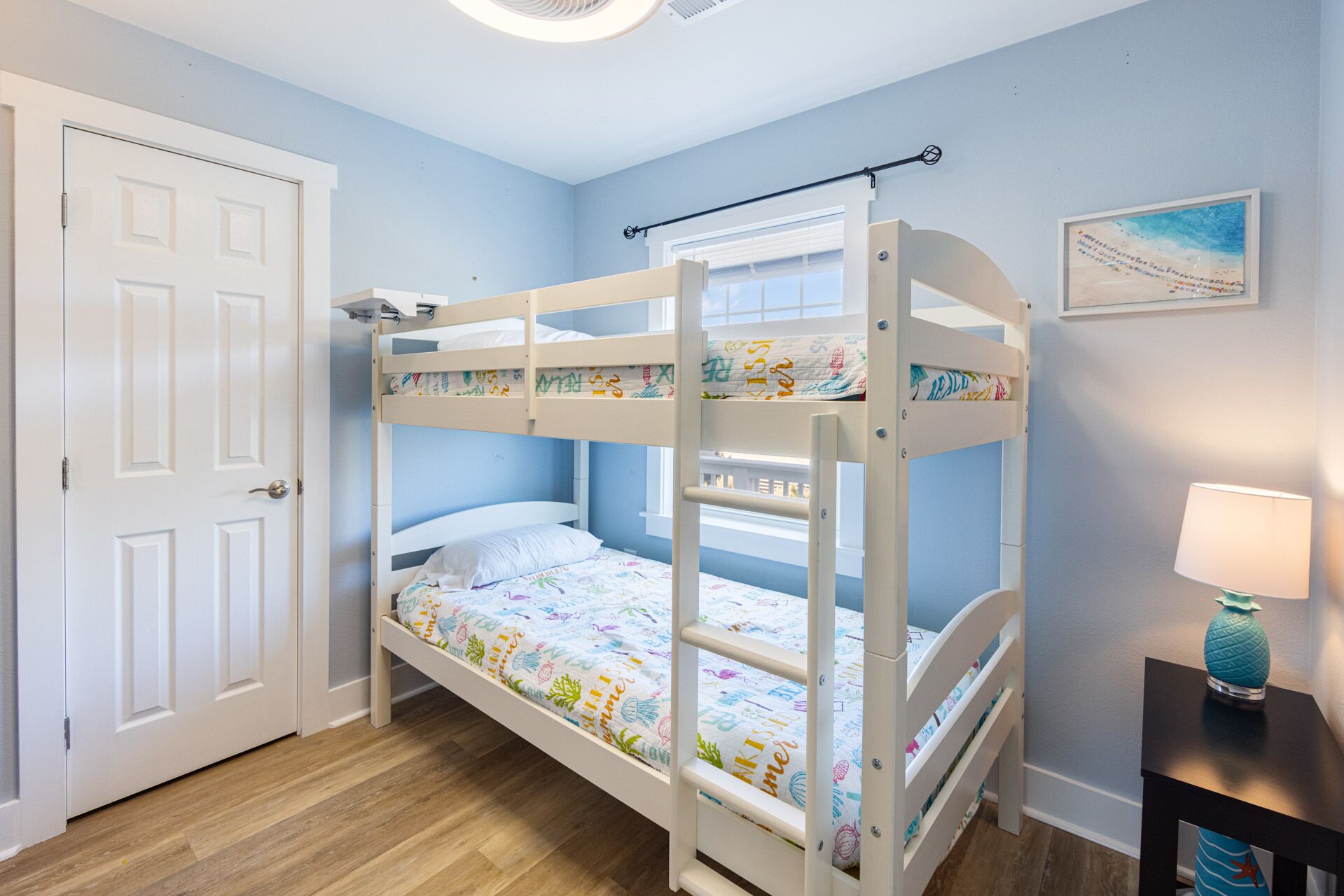 A bunk bed with colorful bedding, a door, a window, and a small table with a lamp.