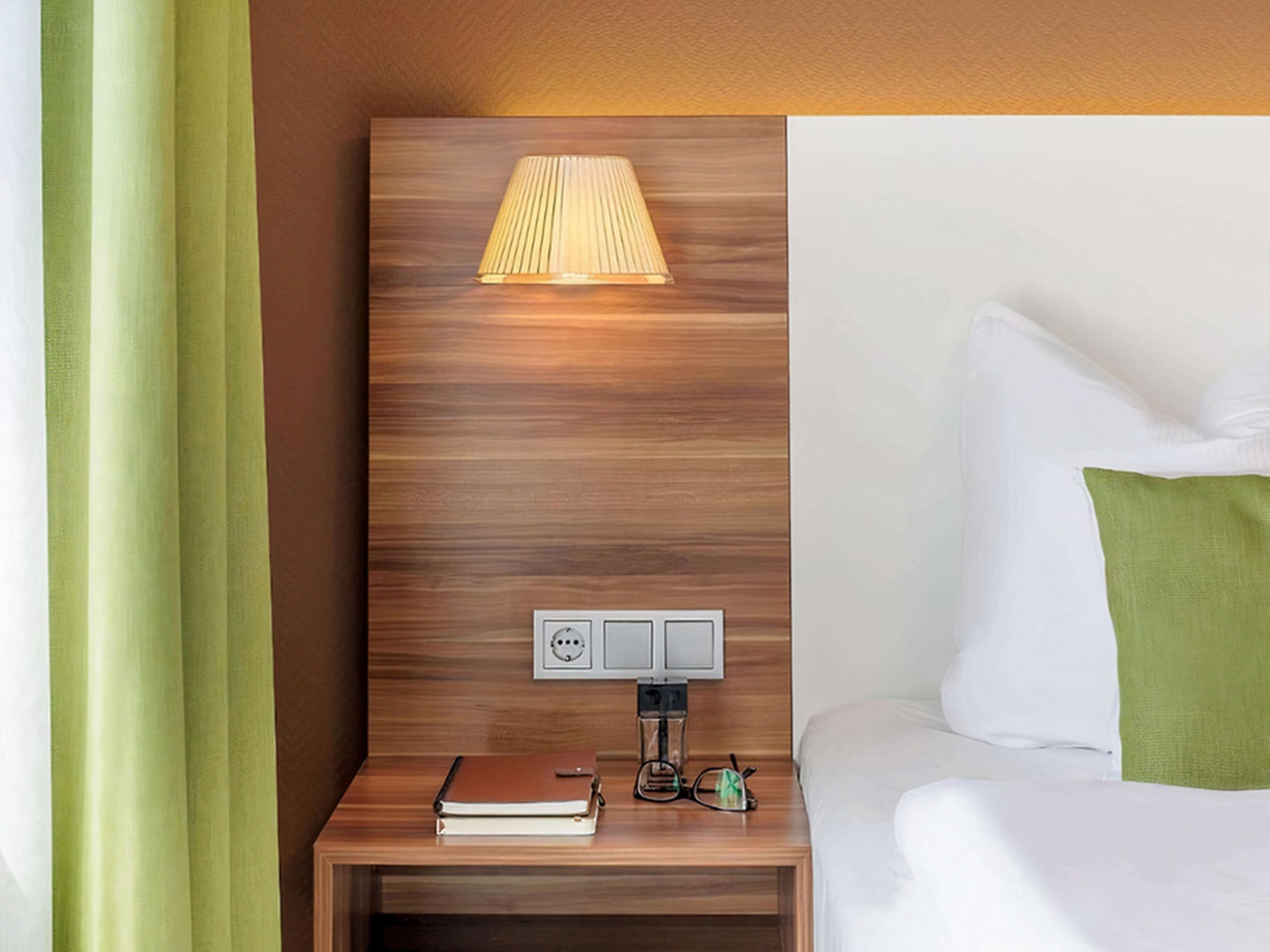 A neatly made bed with white linens and a green pillow, a bedside table with a lamp, a book, and a glass of water.