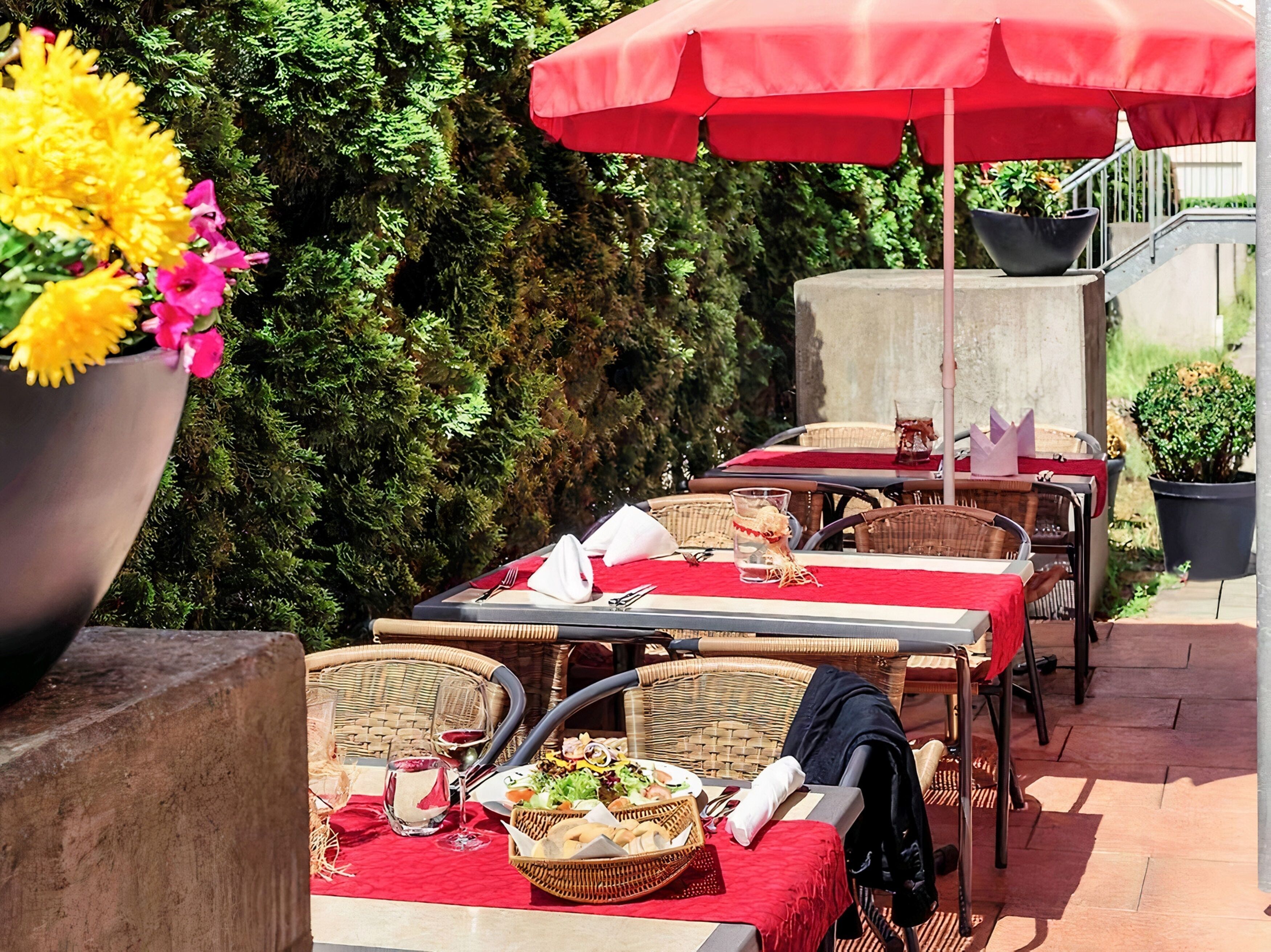 Un área de comedor al aire libre con mobiliario de mimbre, manteles rojos y una planta grande en maceta con flores amarillas.