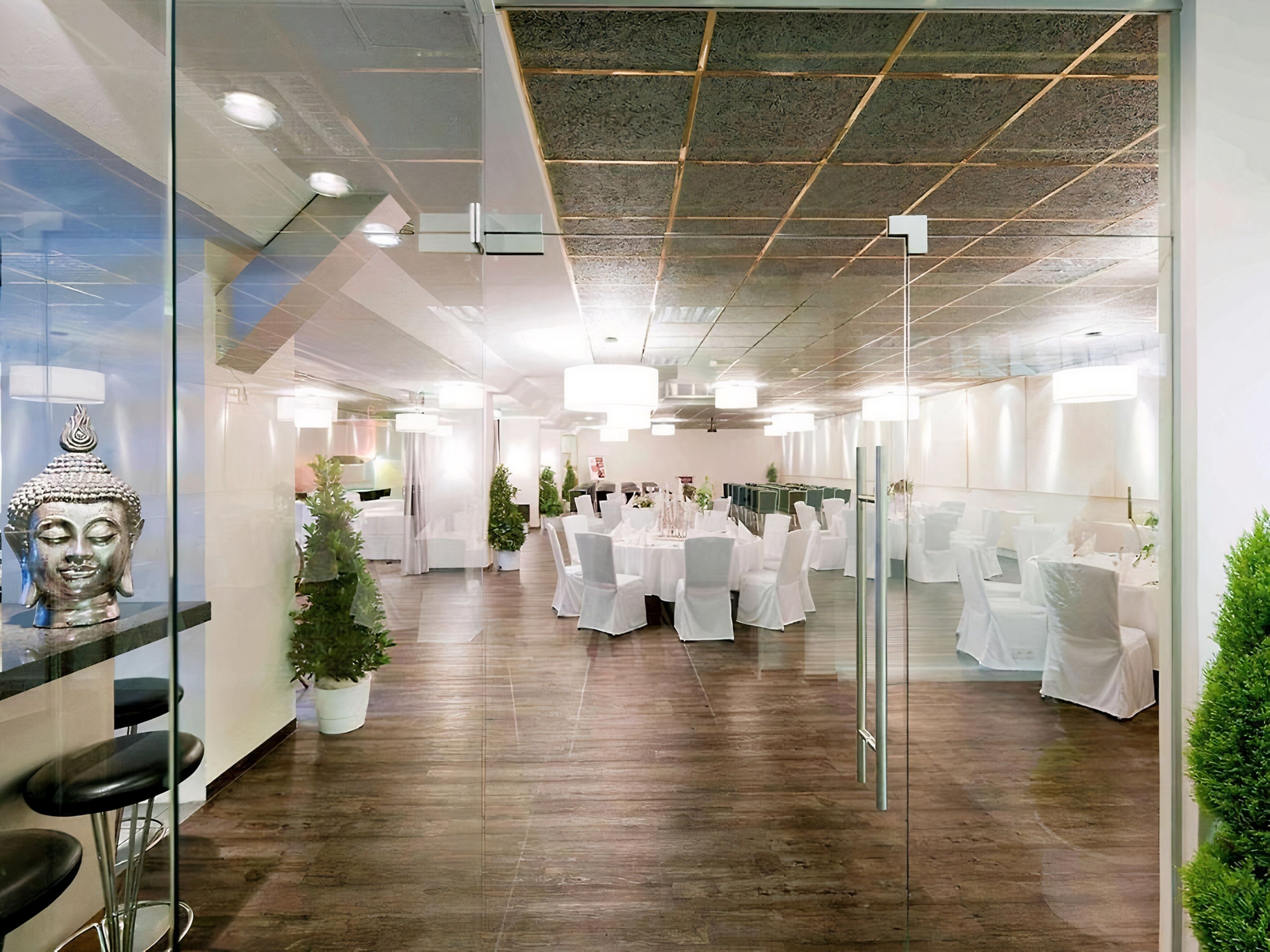 A modern conference room with glass partitions, white tables, and chairs, featuring a Buddha statue and potted plants.