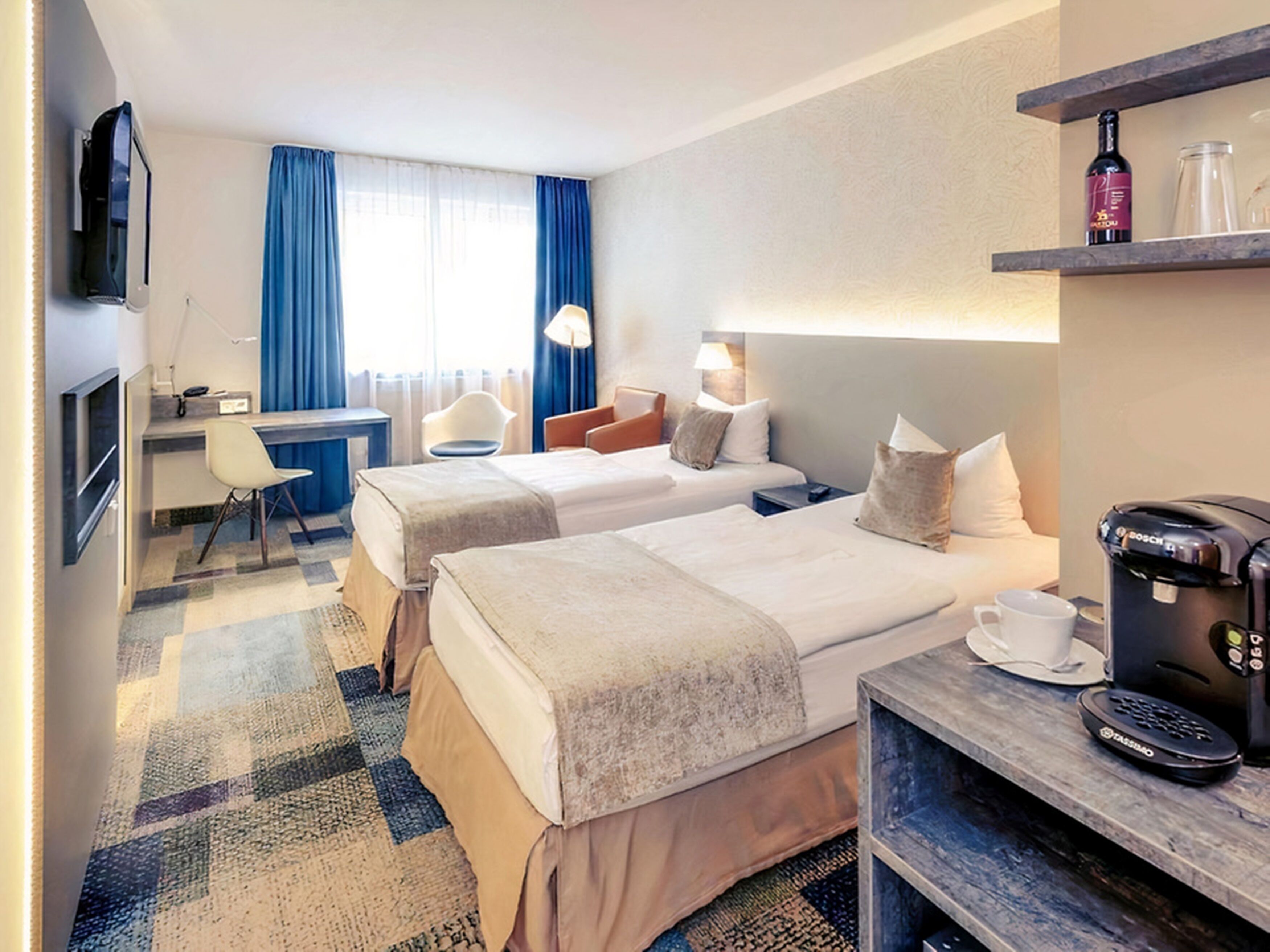 A hotel room with two beds, a desk, a chair, a coffee machine, and a TV.