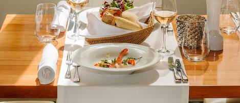 A dining table set with a white tablecloth, a basket of bread, a plate of shrimp, and wine glasses.