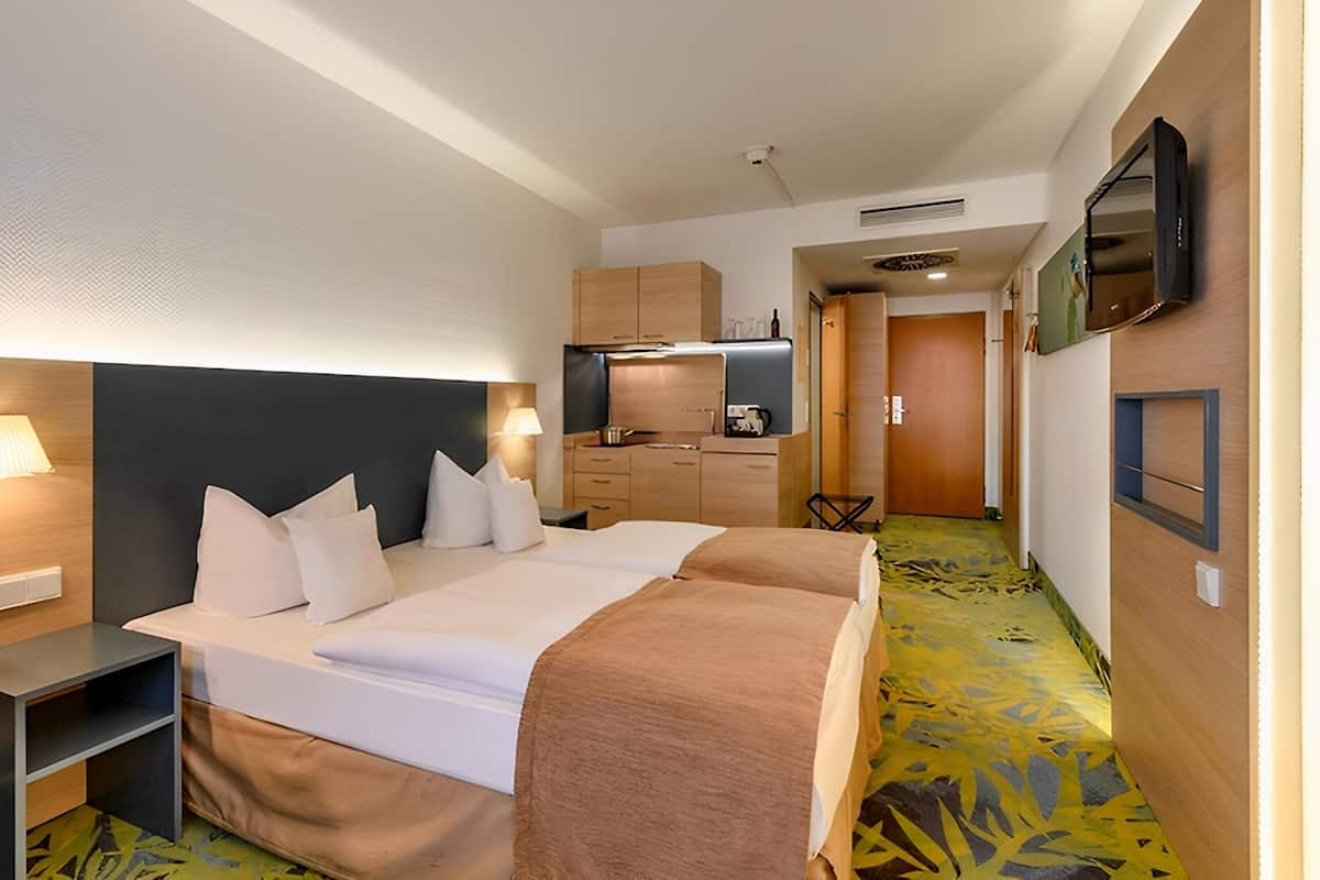 Ein Hotelzimmer mit zwei Betten, einer Küchenzeile, einem an der Wand montierten Fernseher und einem gemusterten Teppich.