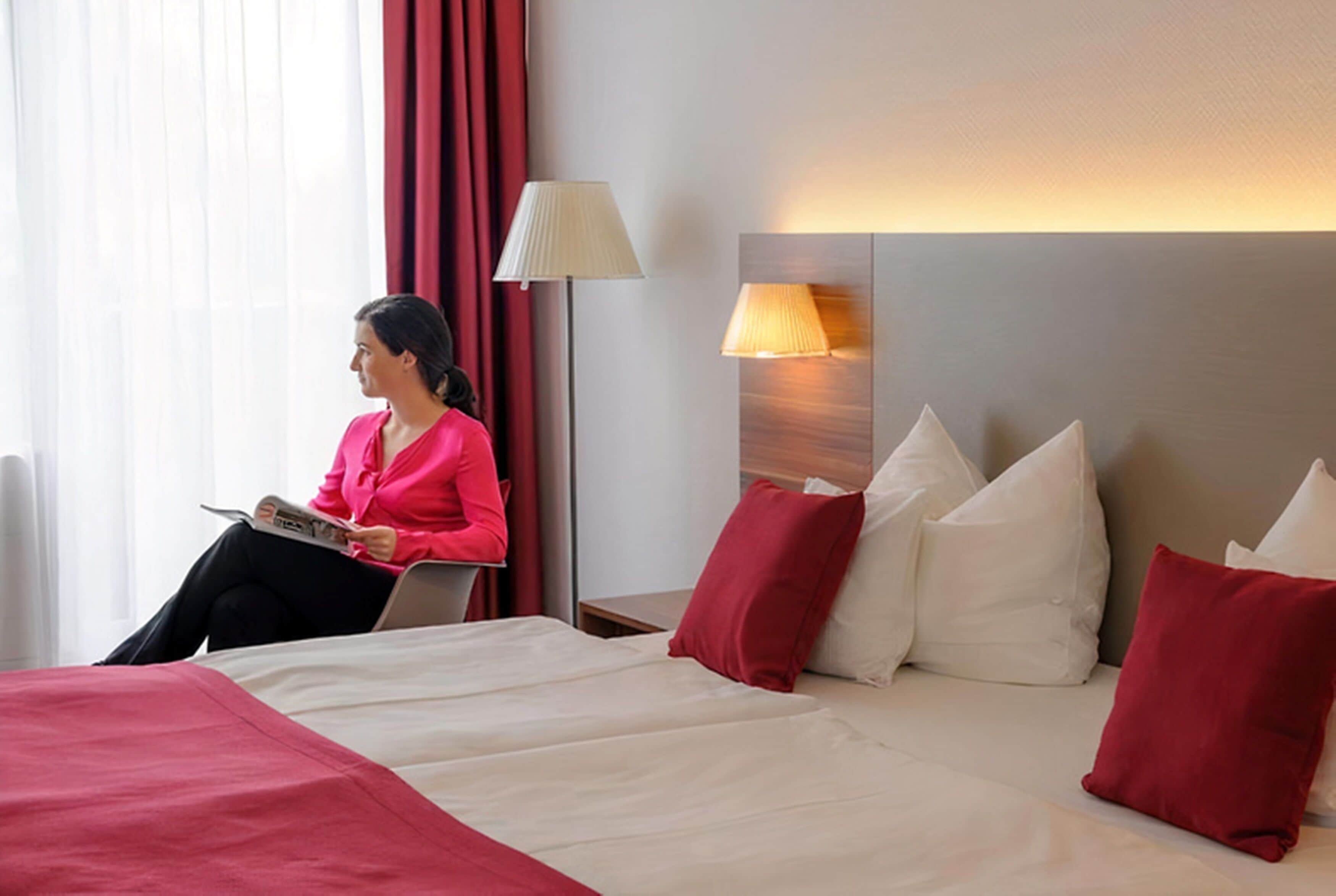 A hotel room with a bed, pillows, a lamp, and a person reading a book.