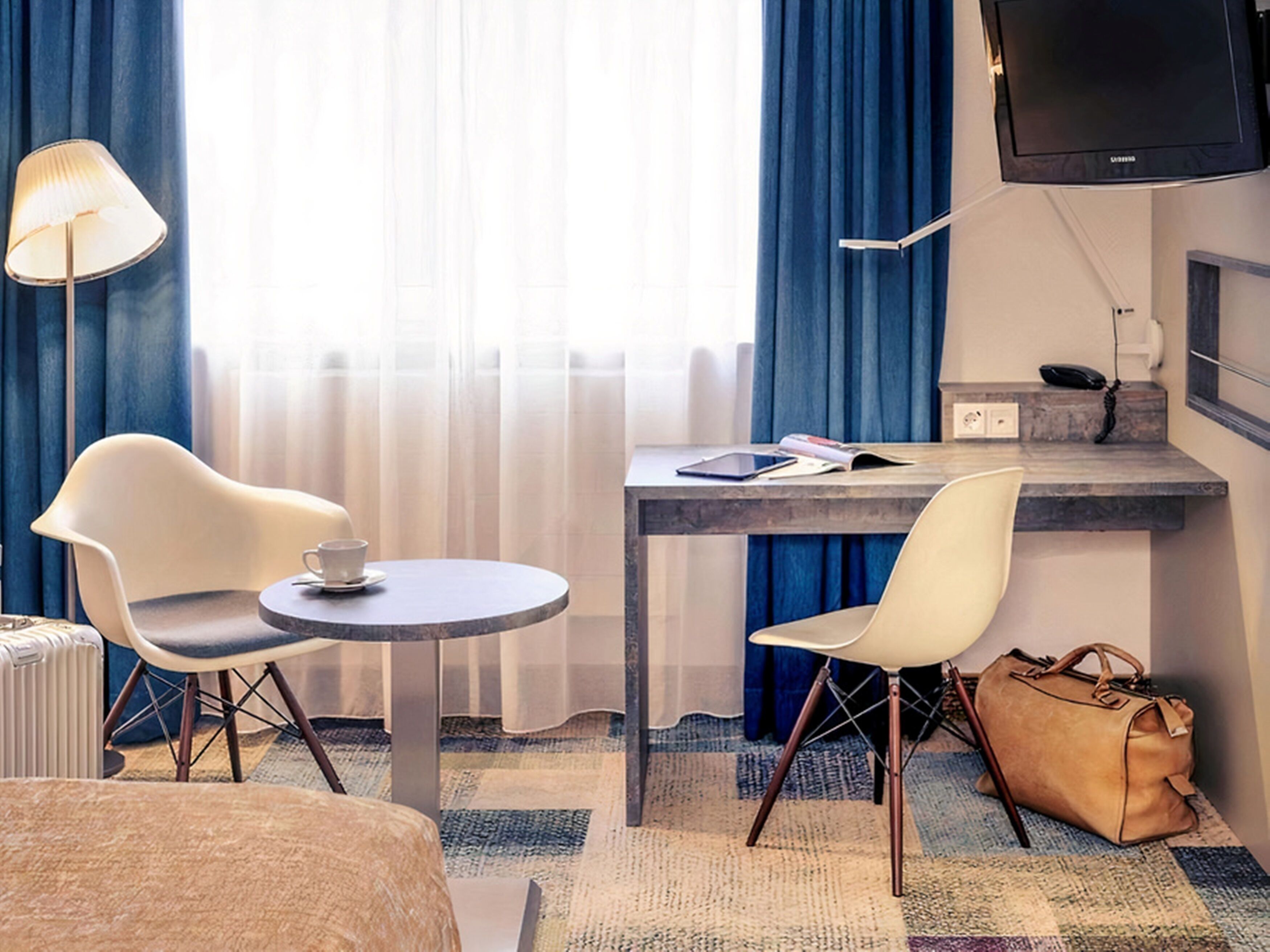 A hotel room with a bed, a desk with a chair, a lamp, a television, and a window with curtains.