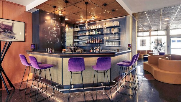 A bar area with a central counter, purple bar stools, and shelves stocked with bottles and glasses.