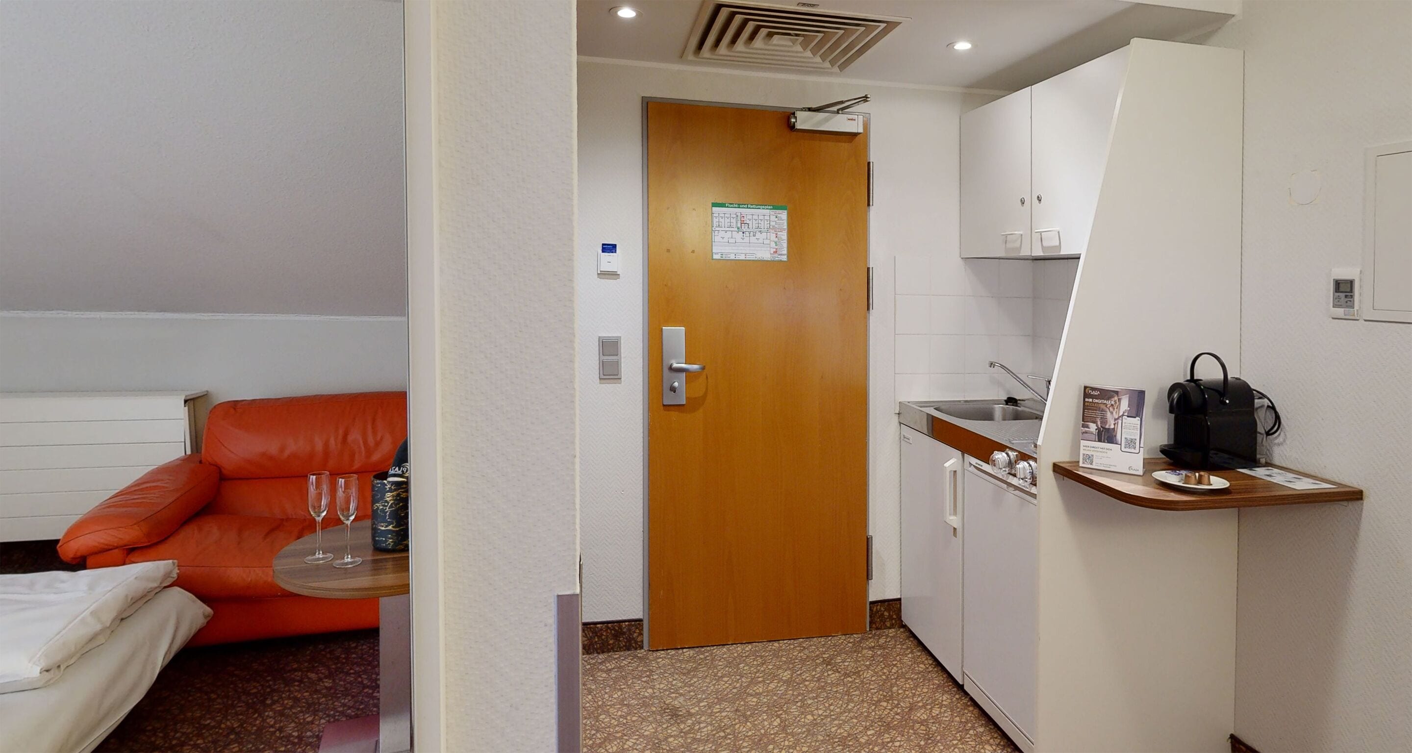 Habitación de hotel compacta con kitchenette, un pequeño comedor con una mesa y dos sillas, y una cama individual con almohada blanca.