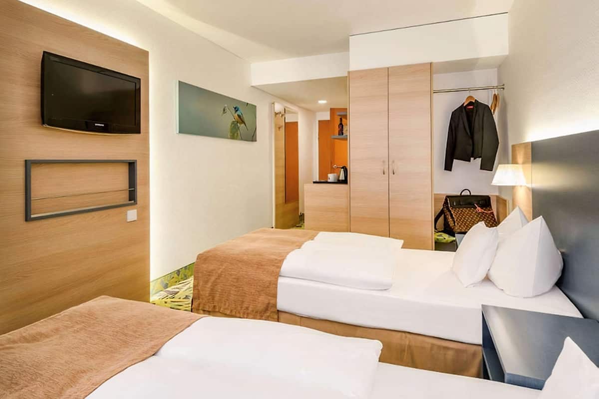 Ein Hotelzimmer mit zwei Betten, einem Fernseher, einem Gemälde, einem Kleiderschrank und einem Kleiderständer.