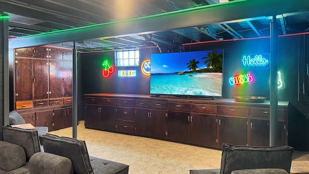 Zona de bar moderna amb un televisor de grans dimensions que mostra una escena de platja, mobles de fusta, cartells de neó i seients còmodes.