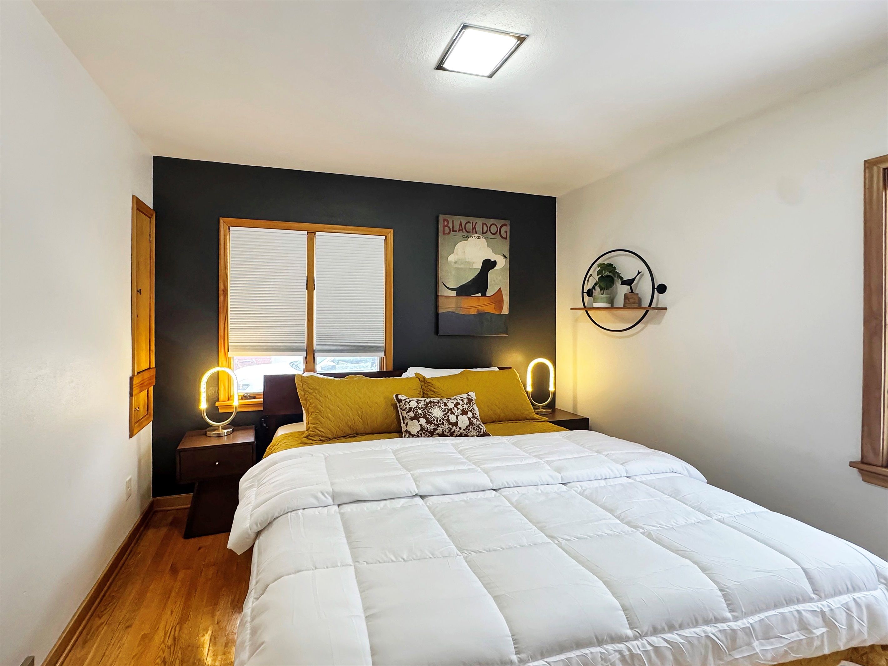 A bedroom with a large bed, a window with blinds, a wall-mounted shelf with decorative items, and a poster of a dog on the wall.
