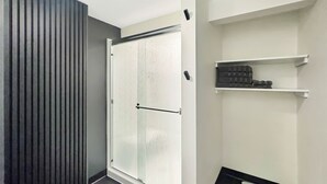 Un bany modern amb porta de dutxa de vidre, terra fosca i paret blanca amb una estant on hi ha productes de tocador.