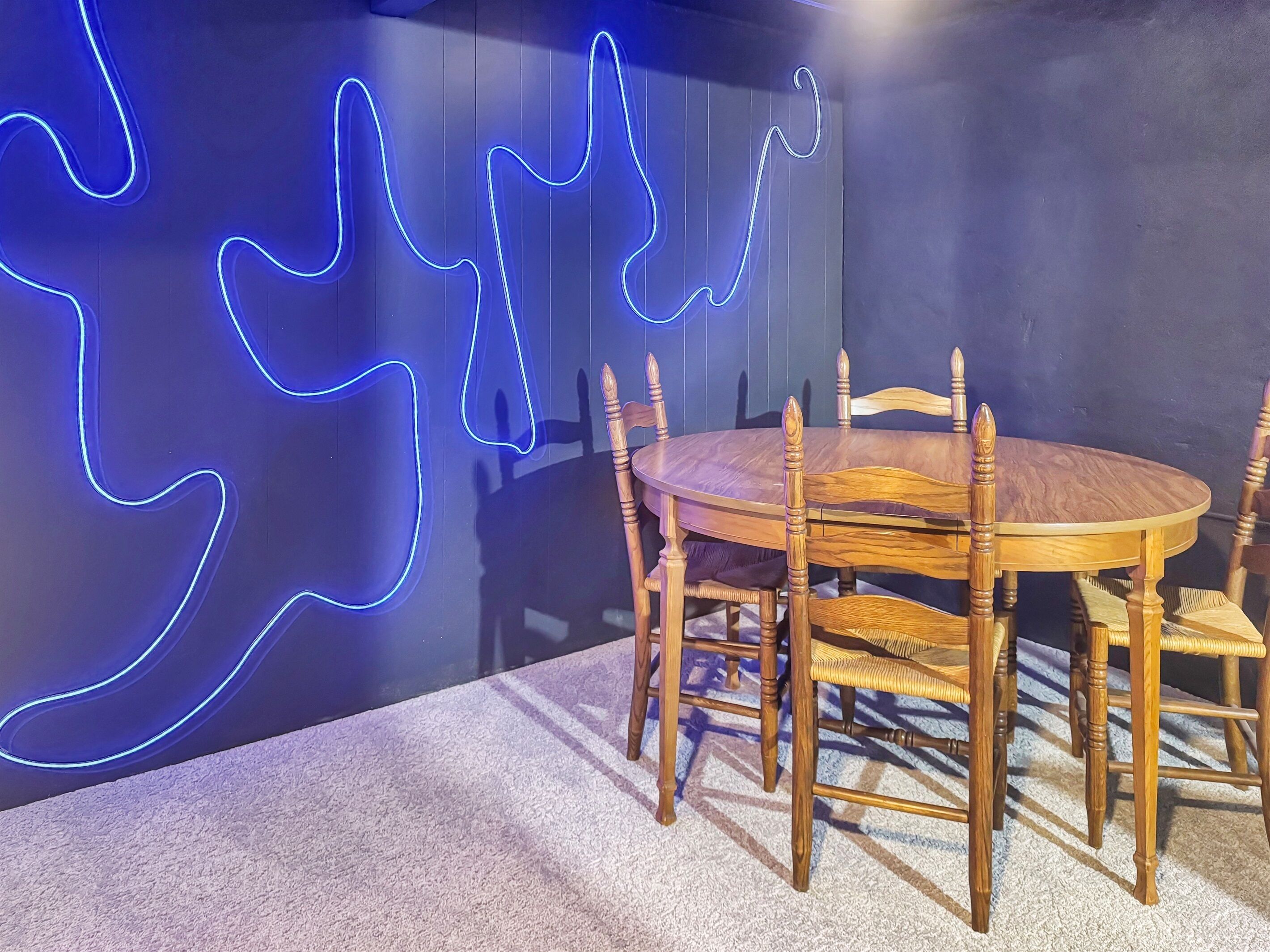 Une table ronde en bois, entourée de six chaises, un mur bleu foncé agrémenté d'un motif lumineux au néon et un sol recouvert d'une moquette.