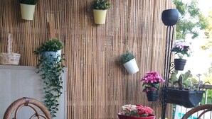 Pared de bambú con plantas en macetas, sillas de mimbre con cojines verdes y una mesa de vidrio.