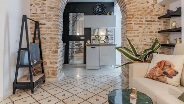 Een woonkamer met een bank, een salontafel, een hondenbedje en een plant. Door een boog is een keuken zichtbaar.