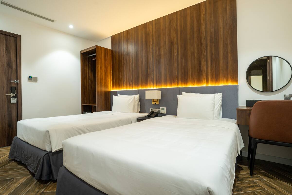 A hotel room with two beds, a wooden accent wall, a round mirror, and a desk with a chair.
