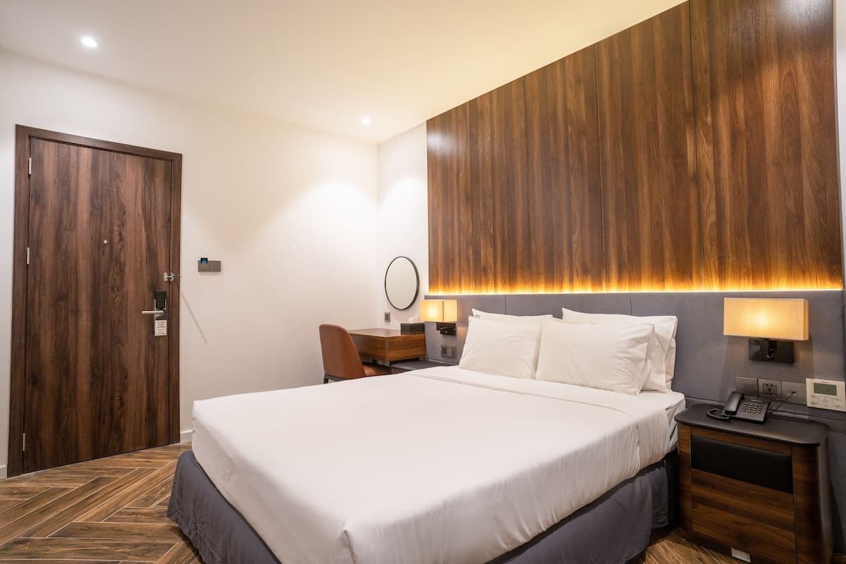 A hotel room with a large bed, a desk, a chair, a bedside table with a lamp, a wall-mounted mirror, and a wooden door.