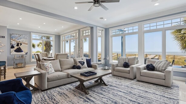 A spacious living room with a large window overlooking a beach, a ceiling fan, and a modern light fixture. - 7 Singleton Beach, Oceanfront, Private Pool (Hilton Head Island)