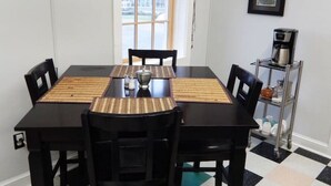 Een eethoek met een zwarte tafel, vier stoelen, een raam met bamboeblinden, een ingelijst beeld aan de muur, en een koffieapparaat op een metalen plank.
