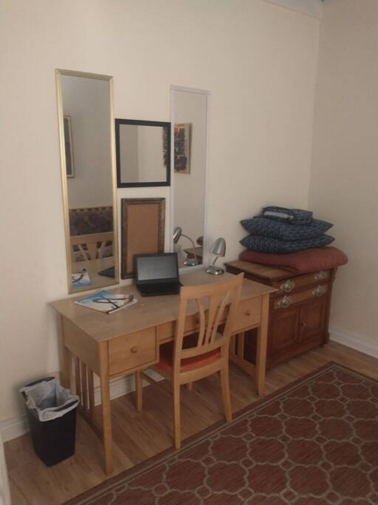 Une petite pièce avec un bureau en bois, une chaise, un ordinateur portable, un miroir, un petit placard et un tapis.