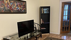 A living room with a glass coffee table, a television, a patterned rug, a bookshelf, and a door.
